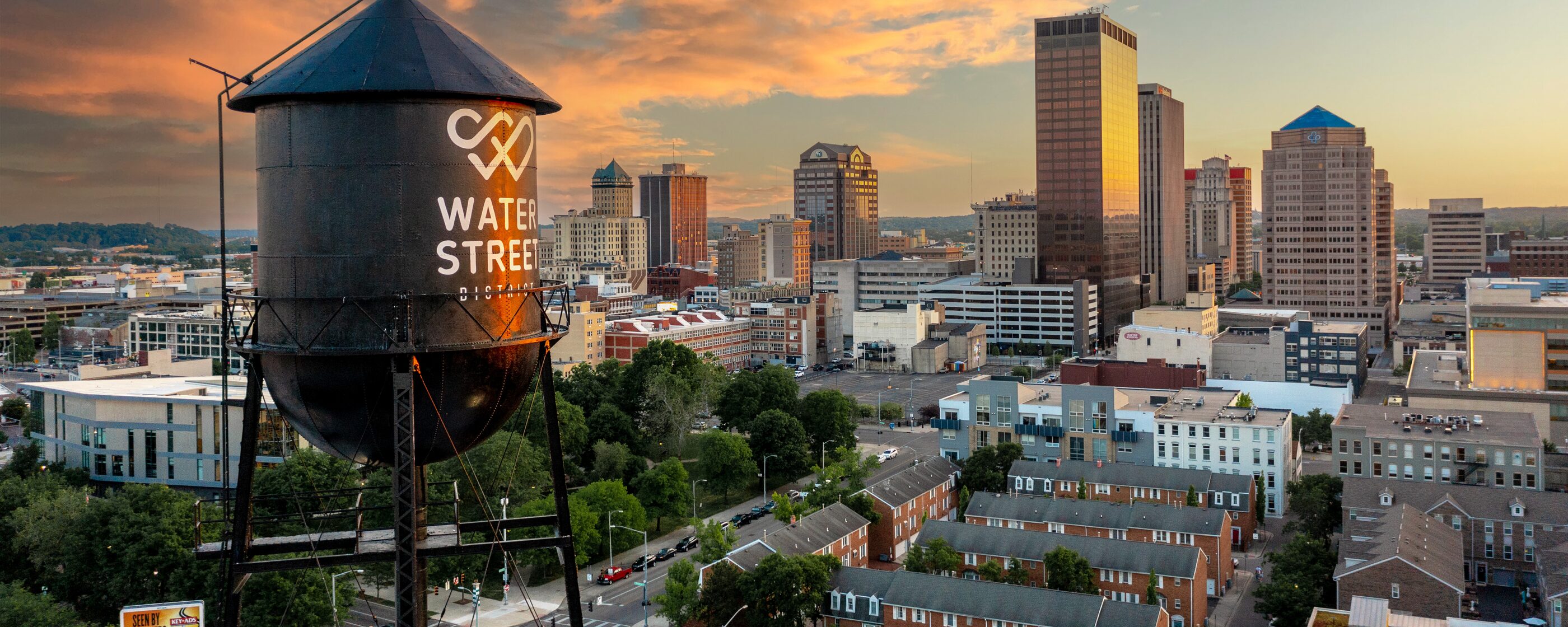 Blocks of Dayton rowhouses and tree-lined streets below the Water Street tower at sunset, downtown behind them