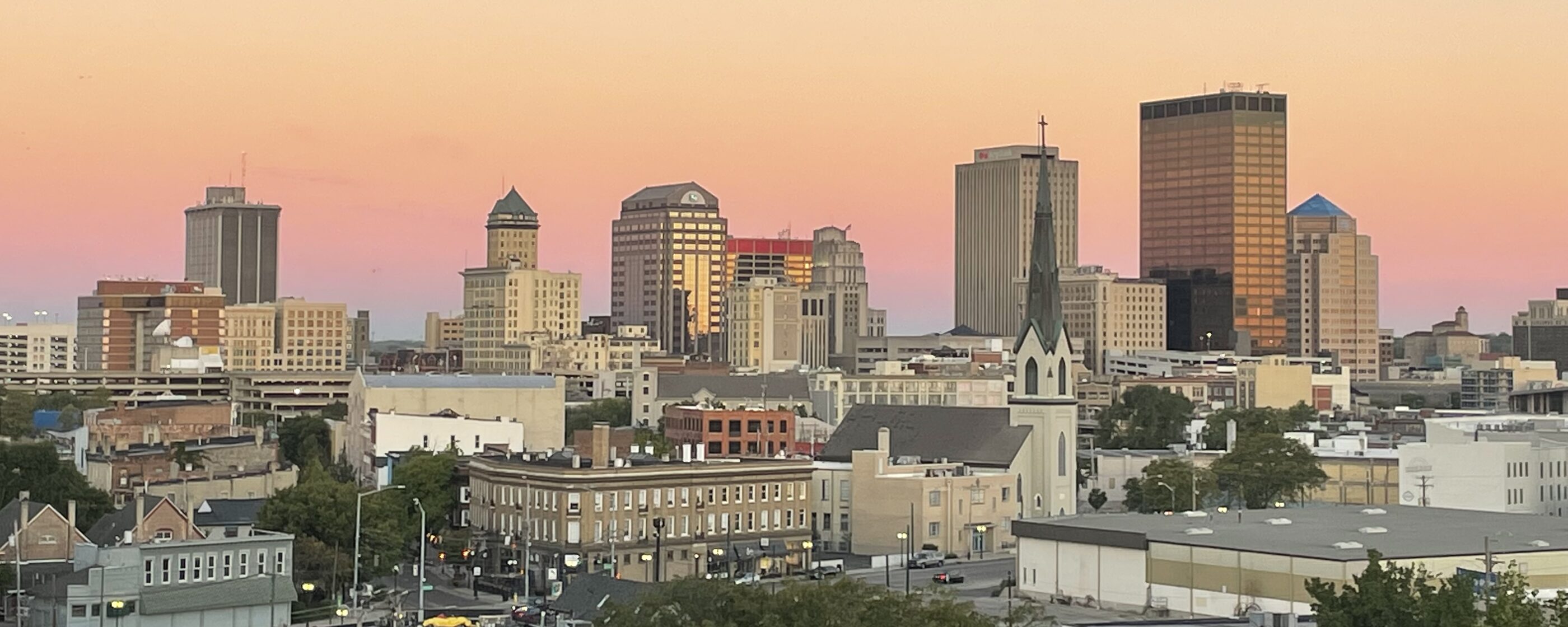 The downtown Dayton skyline under a peach and rose sunset, with rooftops and a church steeple in the foreground